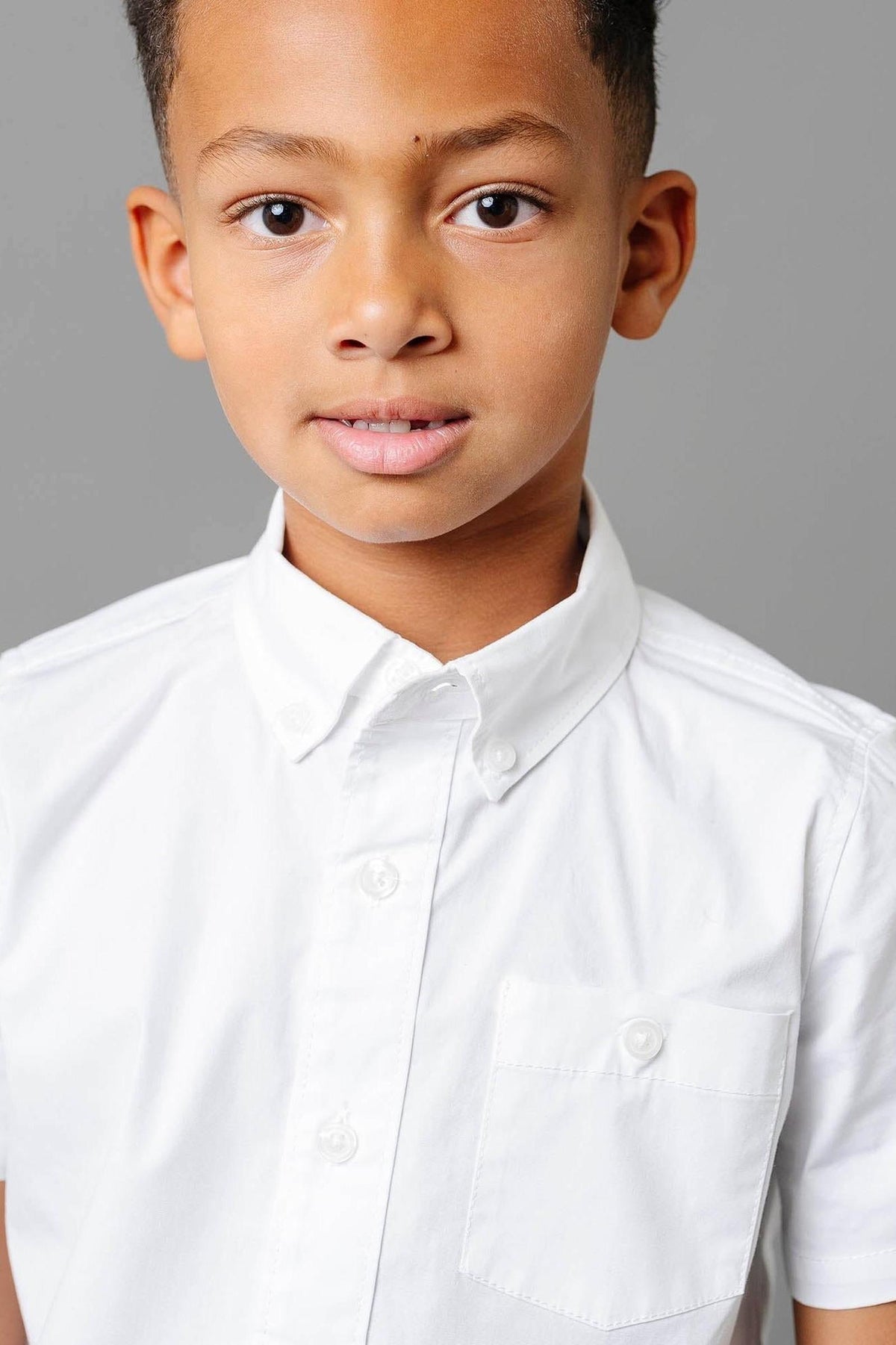 A young boy wearing a white short sleeve button-down shirt with a chest pocket, set against a gray background.