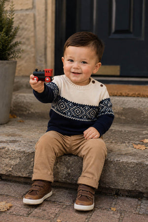 A young boy sits on stone steps, wearing a cream and navy Fair Isle patterned sweater with shoulder buttons. He holds a small red toy train, smiling cheerfully.