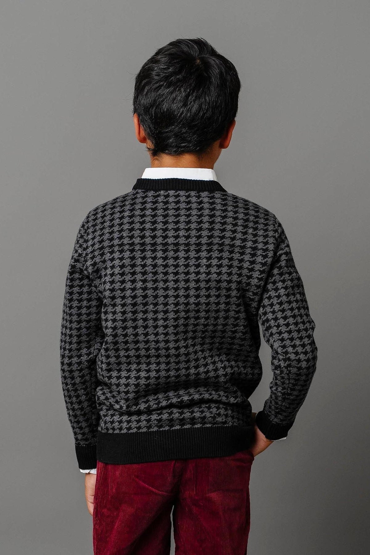 A boy stands with his back facing the camera, wearing a charcoal intarsia knit sweater and burgundy corduroy pants.
