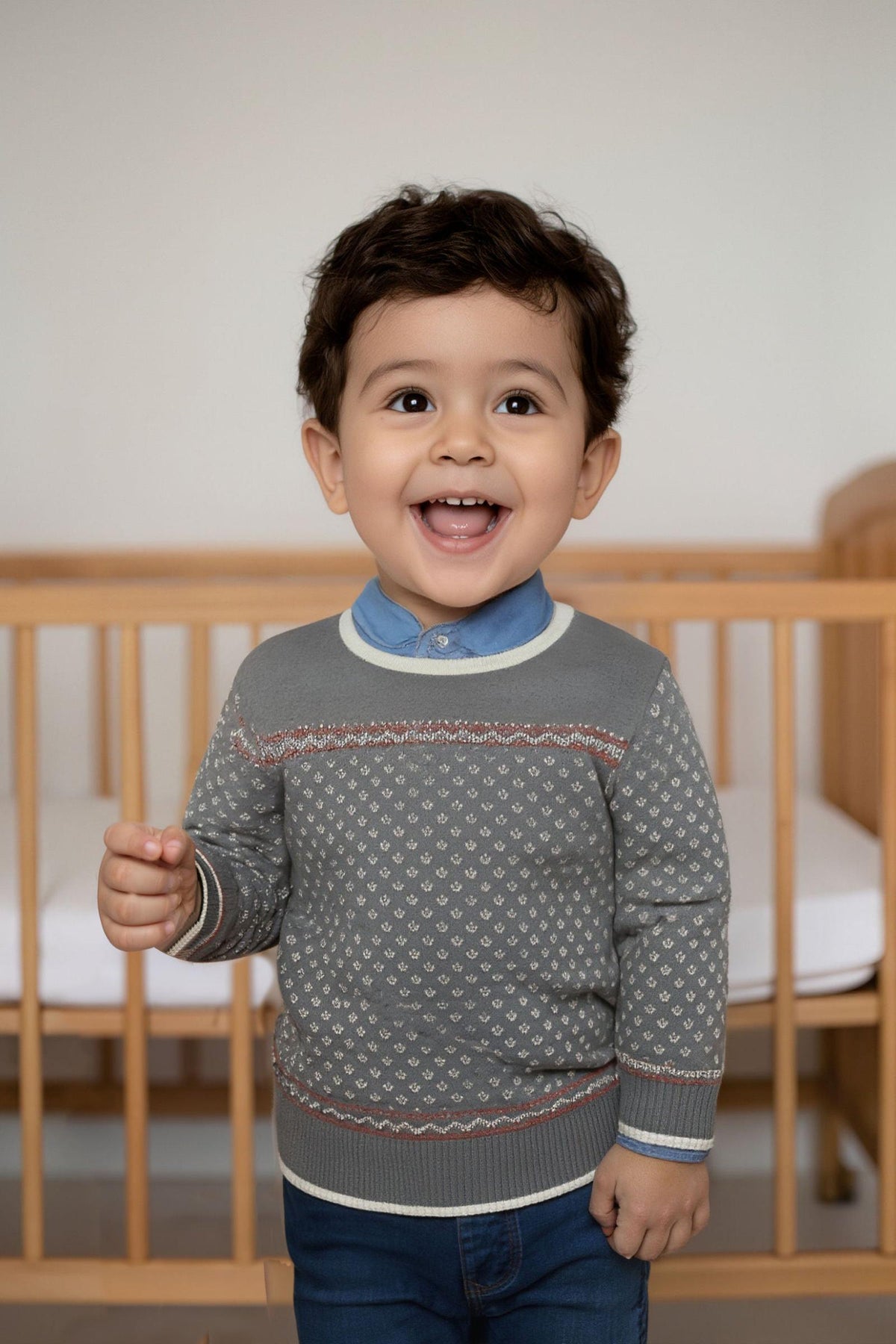 A joyful toddler smiles while wearing a charcoal heather crew neck sweater with an ivory birdseye pattern. The setting features a wooden crib, emphasizing a cozy, family-friendly environment.