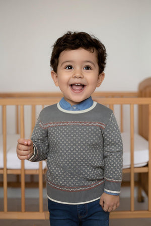 A joyful toddler smiles while wearing a charcoal heather crew neck sweater with an ivory birdseye pattern. The setting features a wooden crib, emphasizing a cozy, family-friendly environment.
