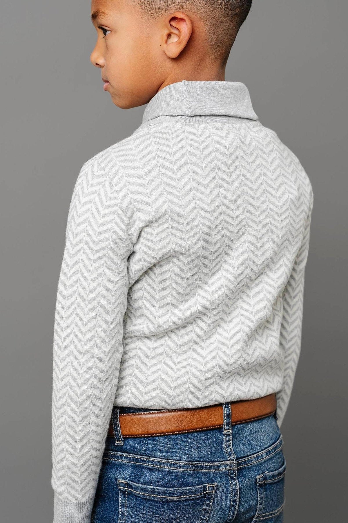 A boy is wearing a grey herringbone knit sweater with a shawl collar, paired with blue jeans and a brown.
