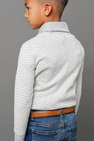 A boy is wearing a grey herringbone knit sweater with a shawl collar, paired with blue jeans and a brown.