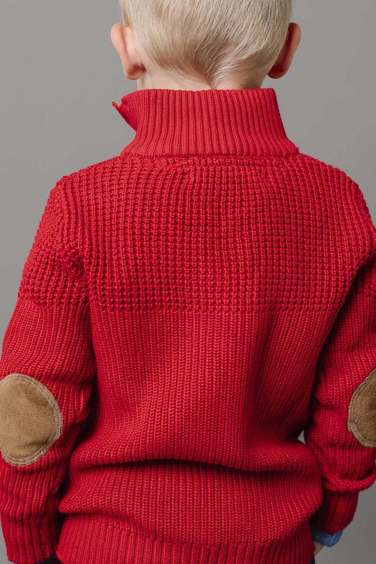 A baby boy is seen from behind wearing a vibrant red mock neck sweater featuring a mixed stitch design and faux suede elbow patches.