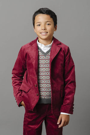 A boy wearing a red corduroy suit and patterned sweater, standing against a gray background.