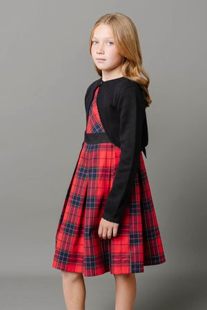 A girl wearing a red tartan pleated dress stands against a gray background, complemented by a black cardigan.
