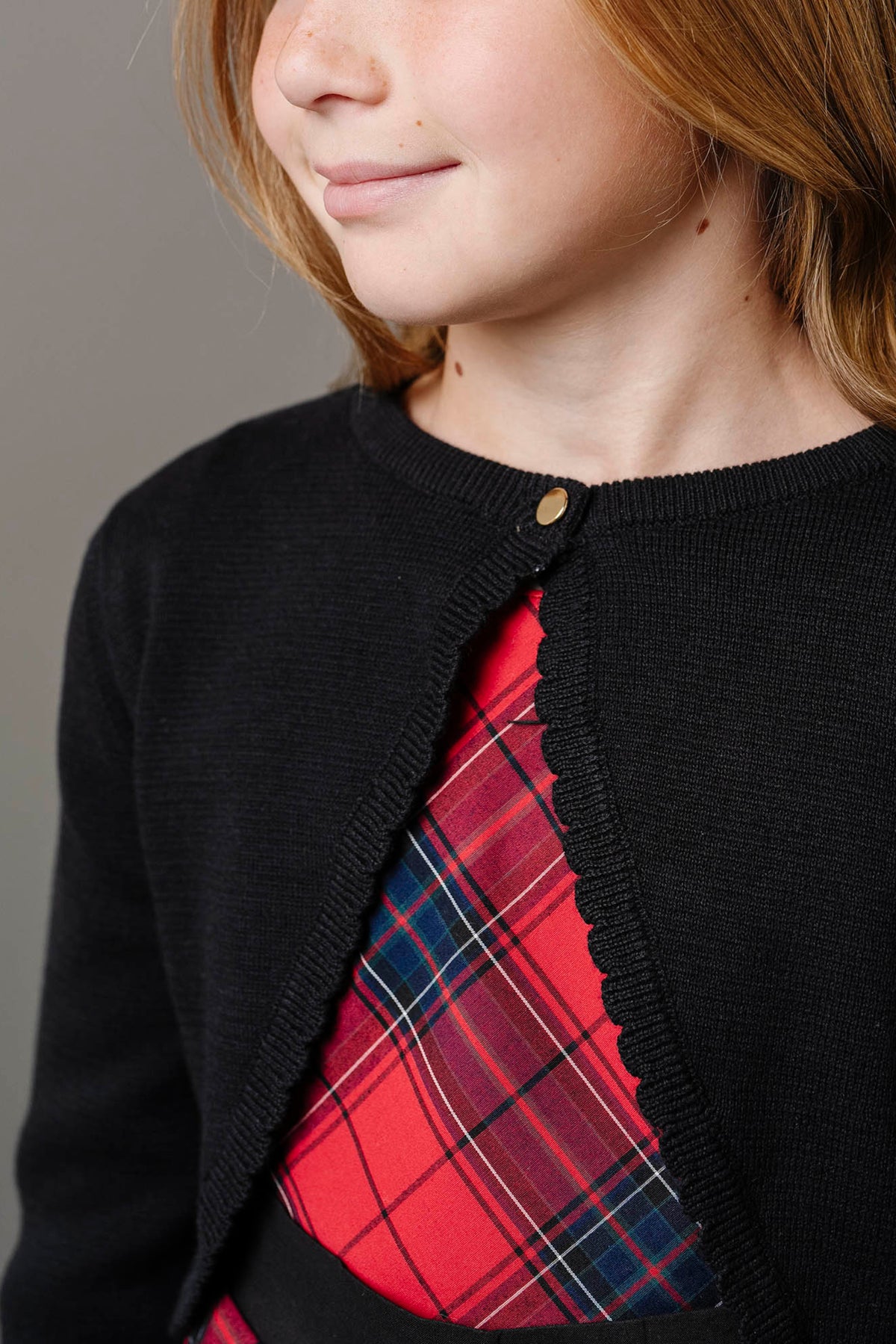 A girl in a black cardigan smiles softly, showcasing a red tartan dress beneath. The cardigan features a scalloped edge and a small gold button, adding a touch of elegance.