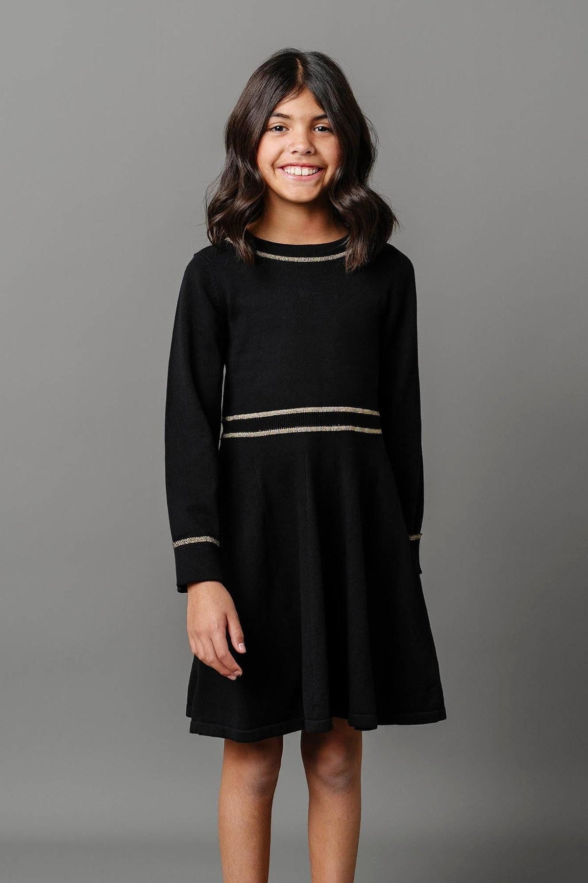 A smiling girl stands wearing a black organic cotton sweater dress with gold tipping at the collar and waist.
