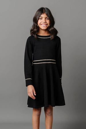 A smiling girl stands wearing a black organic cotton sweater dress with gold tipping at the collar and waist.