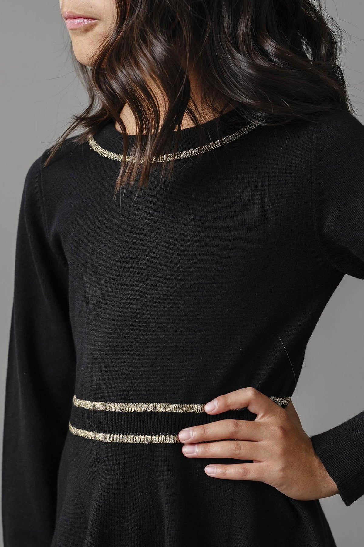 A young child models a black sweater dress featuring gold tipping along the collar and waist. The dress is made from soft organic cotton, perfect for stylish, eco-conscious wear.