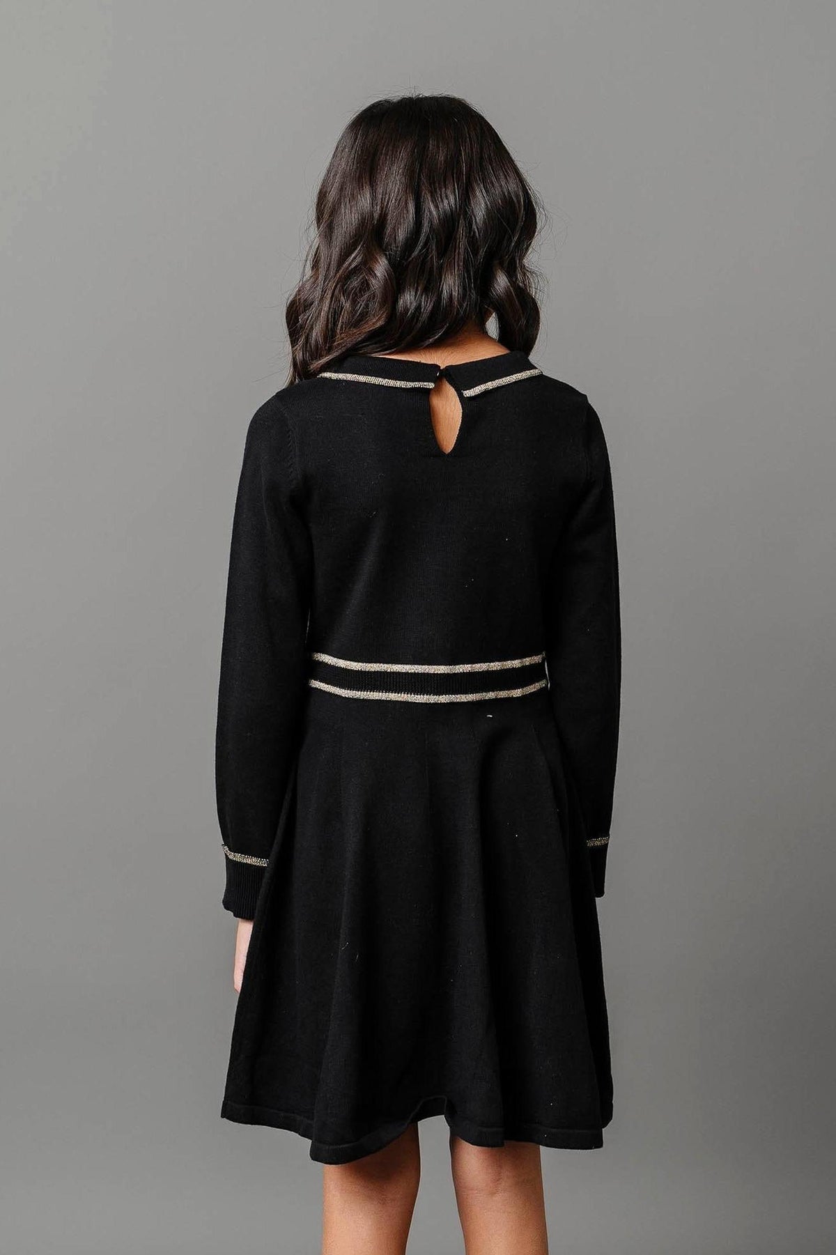 A young girl models a black organic cotton sweater dress featuring gold tipping along the collar and waist. The dress has a keyhole back design, ideal for kids special occasions.