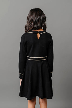 A young girl models a black organic cotton sweater dress featuring gold tipping along the collar and waist. The dress has a keyhole back design, ideal for kids special occasions.