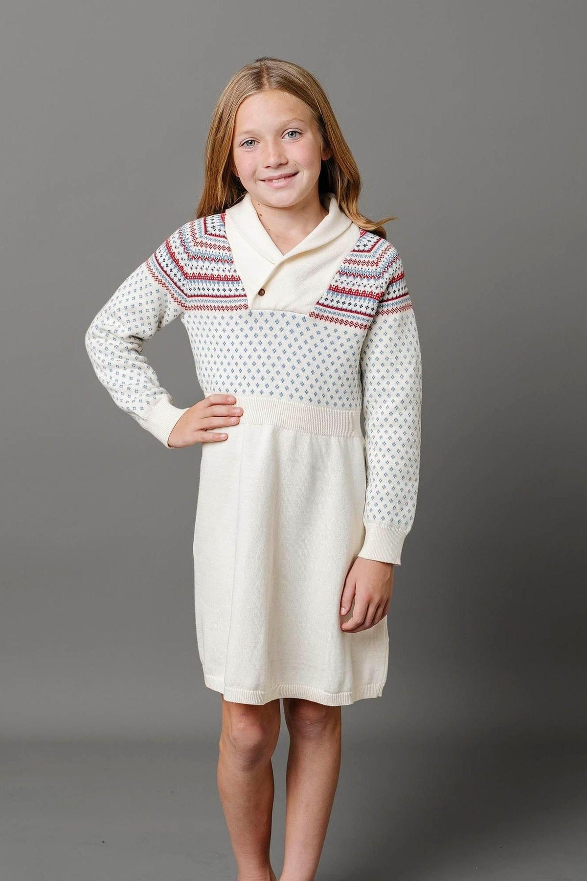 A girl models an ivory organic cotton sweater dress featuring a shawl collar and colorful knit patterns on the bodice.