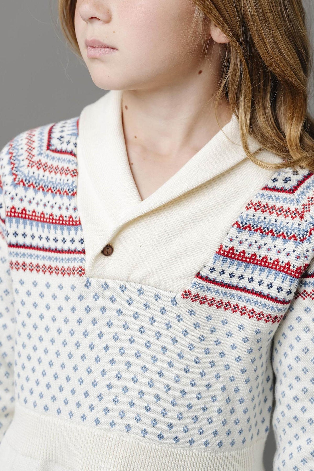 Close-up of a child wearing an ivory organic cotton sweater dress featuring a shawl collar and colorful fair isle patterns.