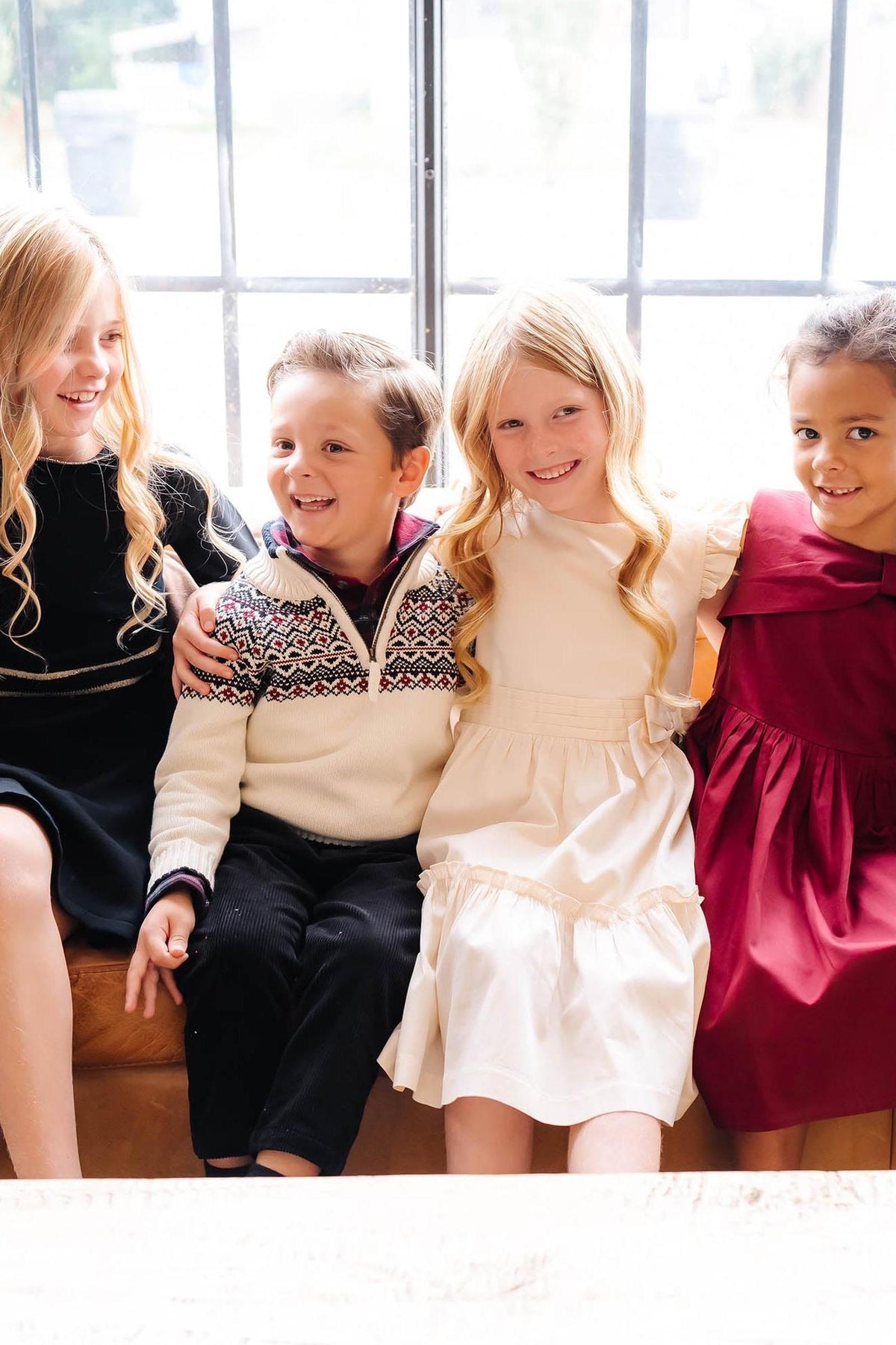 Four children sit together on a wooden bench, dressed in various festive outfits, including a black dress, a cream dress, and a red dress.