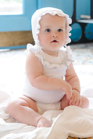 A baby sits on a soft blanket, wearing a white ruffle sweater romper and matching bonnet, showcasing organic cotton material.