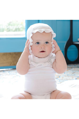 A baby girl sits indoors, wearing a white ruffle romper and matching bonnet, showcasing soft organic cotton material.