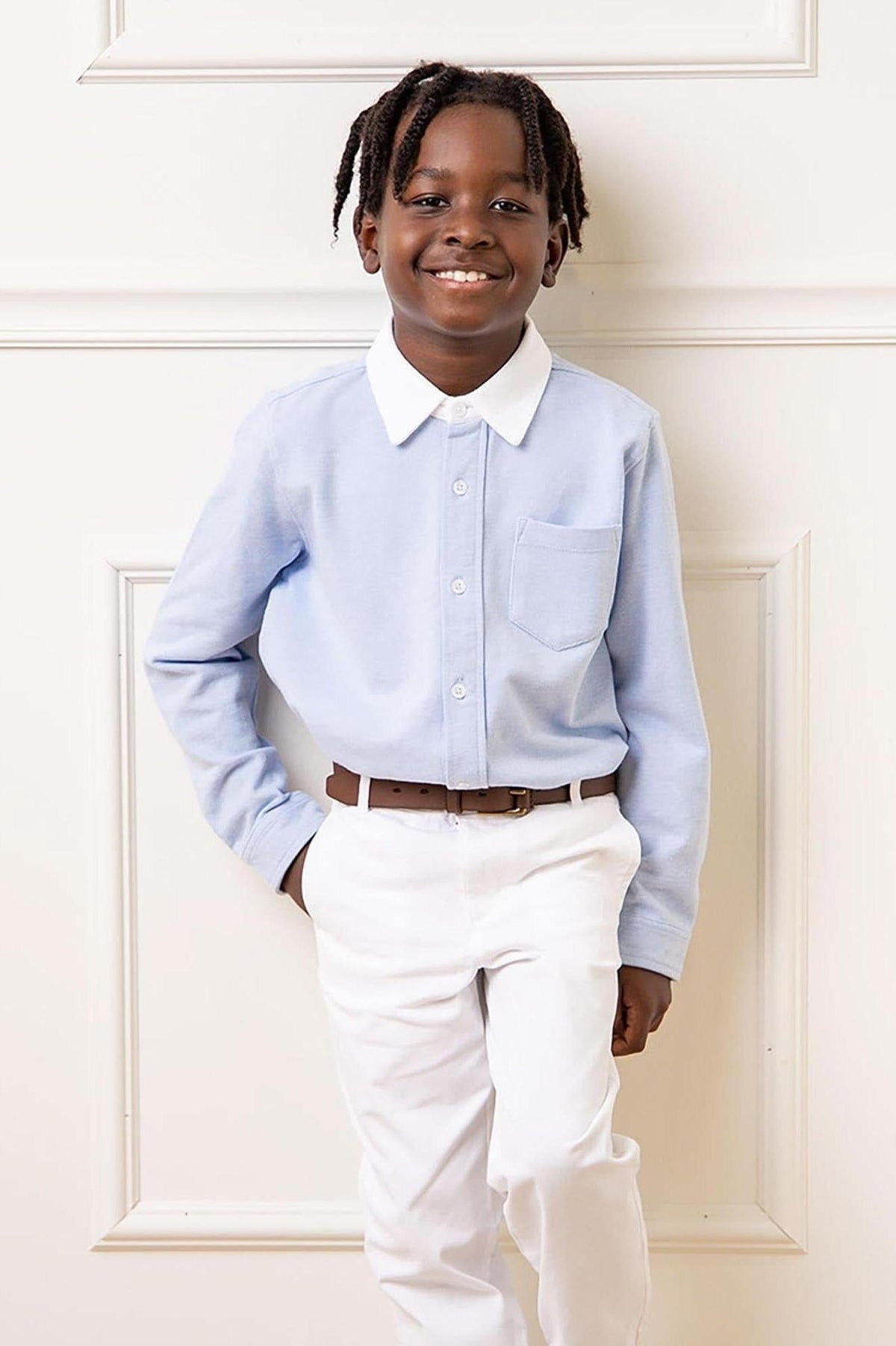 A smiling child models a light blue button-down shirt with a white collar and pocket, paired with white pants. The shirt is made from organic cotton, ideal for ages 2T-10.