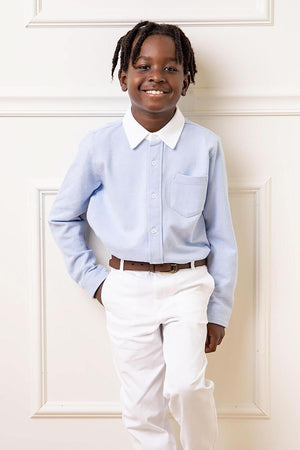 A smiling child models a light blue button-down shirt with a white collar and pocket, paired with white pants. The shirt is made from organic cotton, ideal for ages 2T-10.