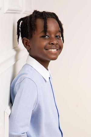 A smiling boy leans against a white wall, wearing a light blue button-down shirt with a white collar. The shirt is made from organic cotton, emphasizing comfort and sustainability.