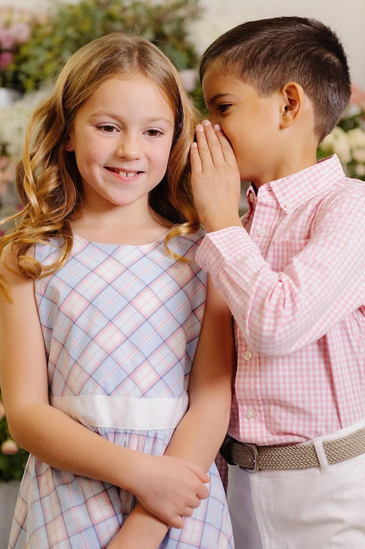 A boy in a pink checkered shirt whispers to a girl wearing a light blue and white checked dress. They are surrounded by a floral backdrop, creating a cheerful atmosphere.