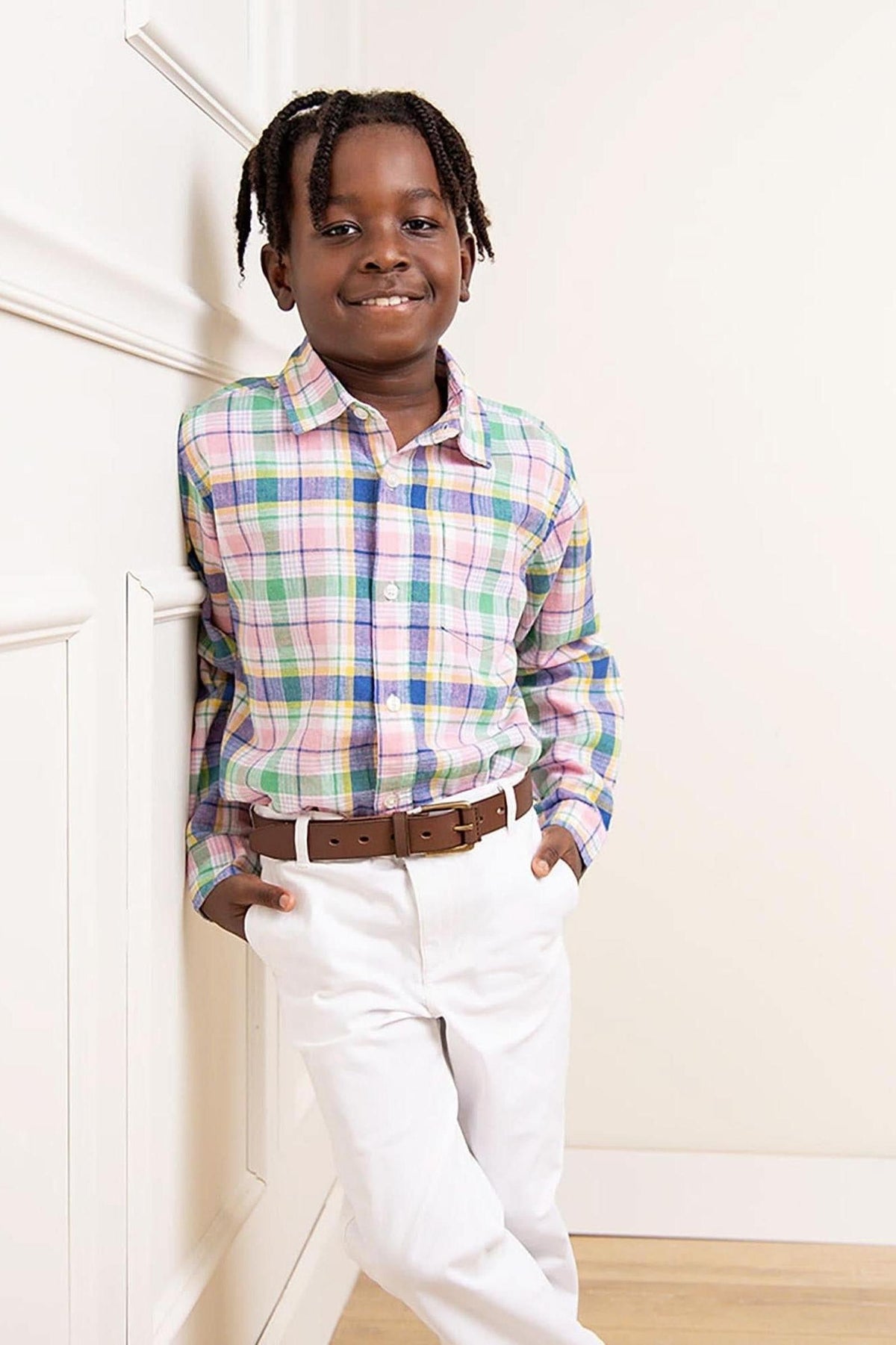 A smiling boy leans against a wall, wearing a colorful plaid button-down shirt and white pants with a brown belt.