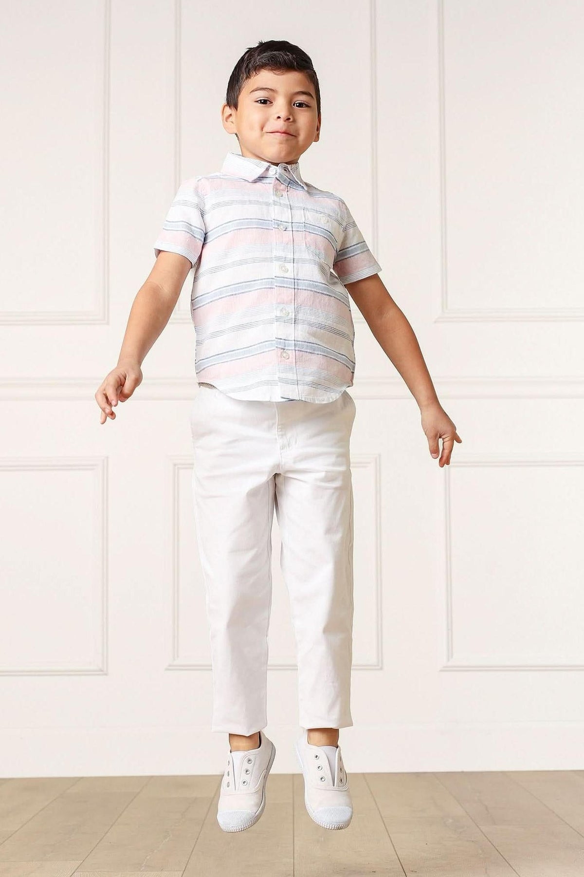 A young boy jumps in the air, wearing a short-sleeve, striped button-down shirt in pastel colors and white pants, showcasing a playful and stylish summer look.