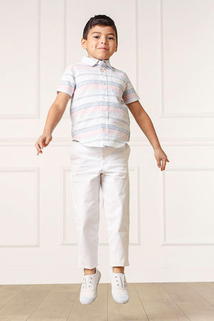 A young boy jumps in the air, wearing a short-sleeve, striped button-down shirt in pastel colors and white pants, showcasing a playful and stylish summer look.