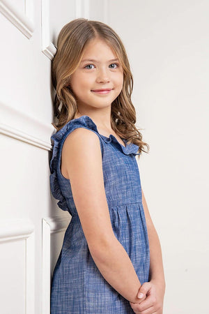 A young girl poses in a chambray organic cotton jumpsuit with ruffle details, against a neutral background.