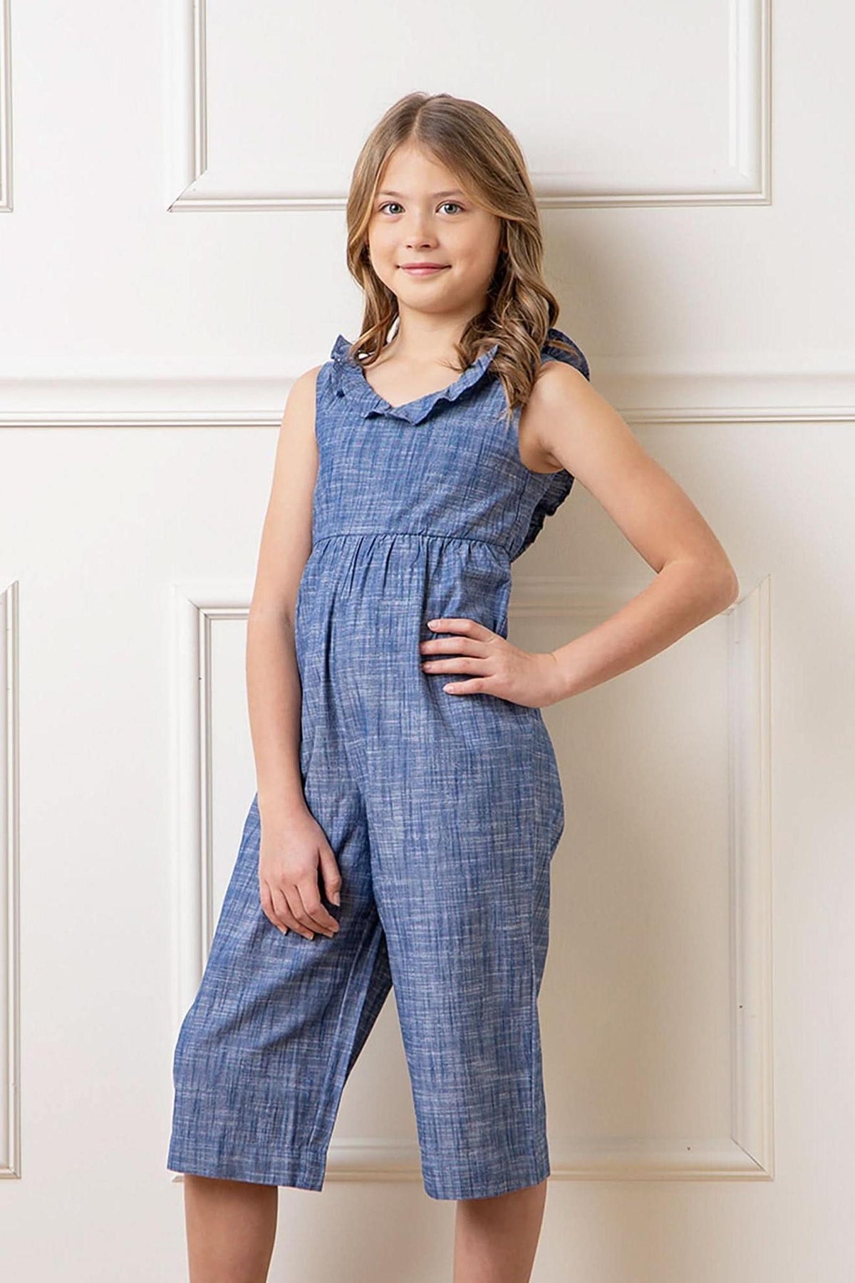 A young girl poses in a chambray organic cotton jumpsuit with a ruffle neck, showcasing a comfortable and stylish design.