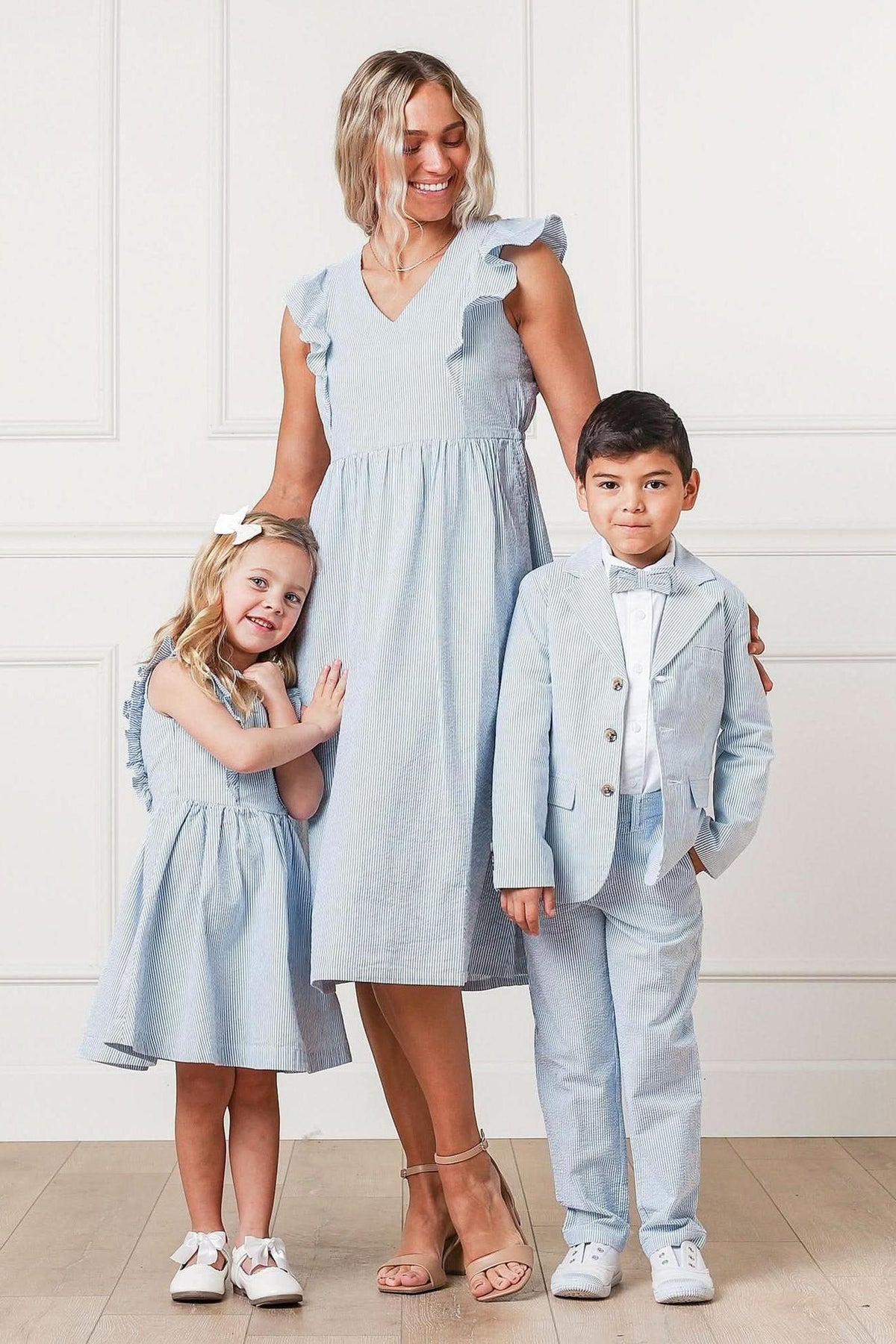 A woman in a light blue dress stands beside a boy in a light blue suit and a girl in a matching dress, all smiling against a white paneled background.