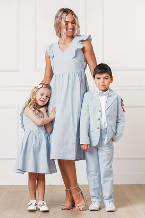 A woman in a light blue dress stands beside a boy in a light blue suit and a girl in a matching dress, all smiling against a white paneled background.