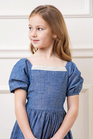 A young girl models a chambray dress with bubble sleeves and a crochet trim, set against a light-colored background.