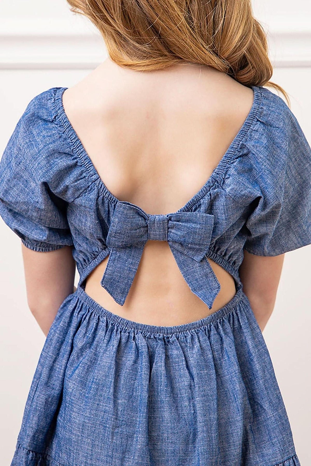 Back view of a chambray dress featuring a bow tie, elastic waistband, and puff sleeves, made from soft organic cotton.