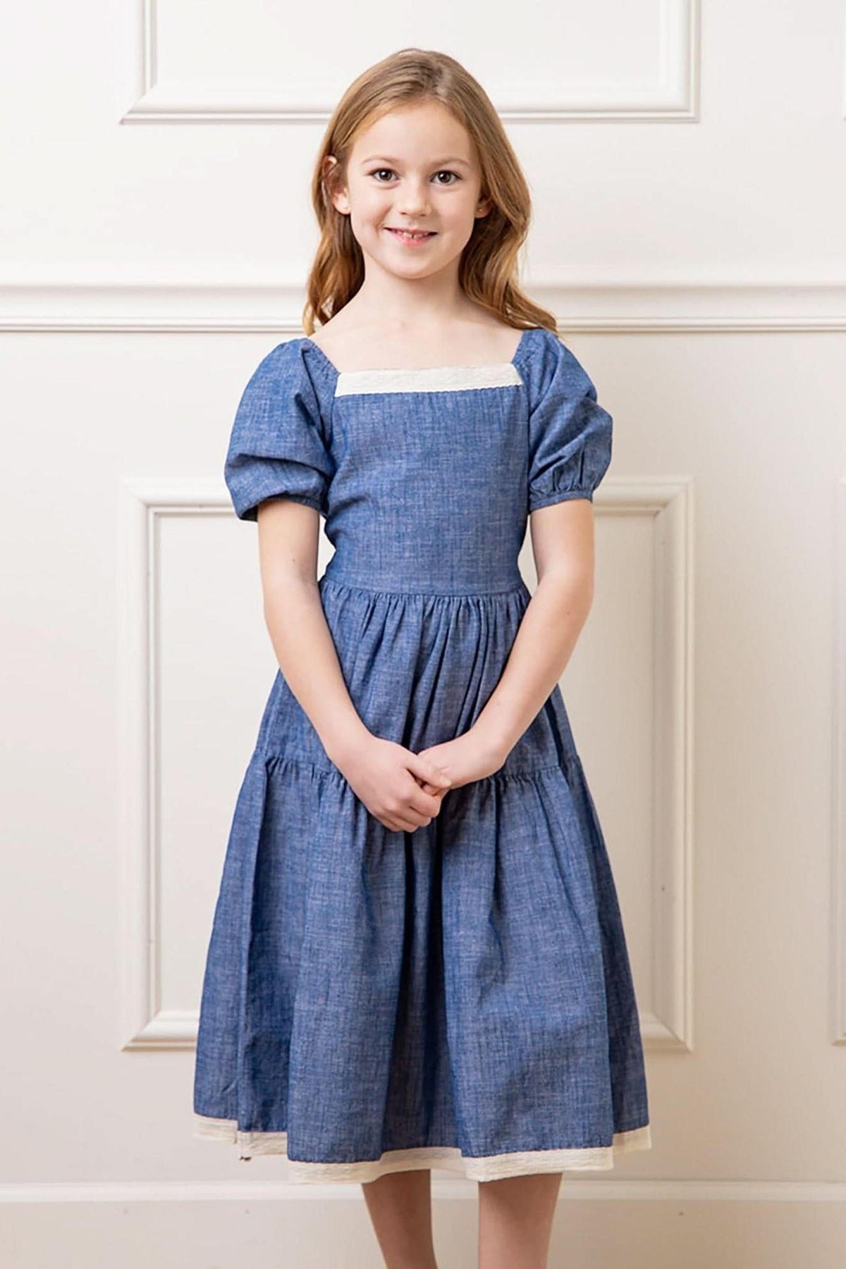 A young girl wears a chambray dress with bubble sleeves and a crochet trim, standing against a light-colored wall.