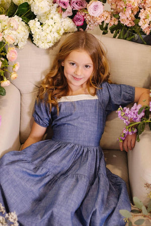 A young girl with wavy hair smiles while wearing a chambray dress, surrounded by colorful flowers on a beige sofa.