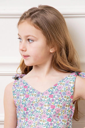 A young girl models a floral dress with bow shoulder straps, showcasing a v-neck silhouette. The fabric features a colorful floral pattern, ideal for summer wear.