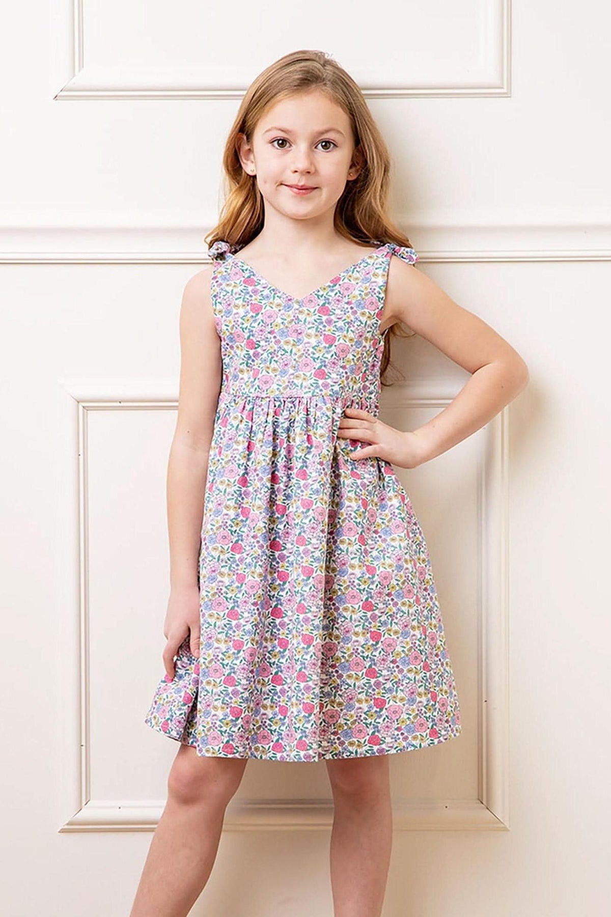A young girl poses in a floral linen dress featuring bow shoulder straps. The dress has a v-neck silhouette and a colorful print, perfect for summer outings.