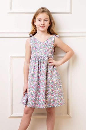 A young girl poses in a floral linen dress featuring bow shoulder straps. The dress has a v-neck silhouette and a colorful print, perfect for summer outings.