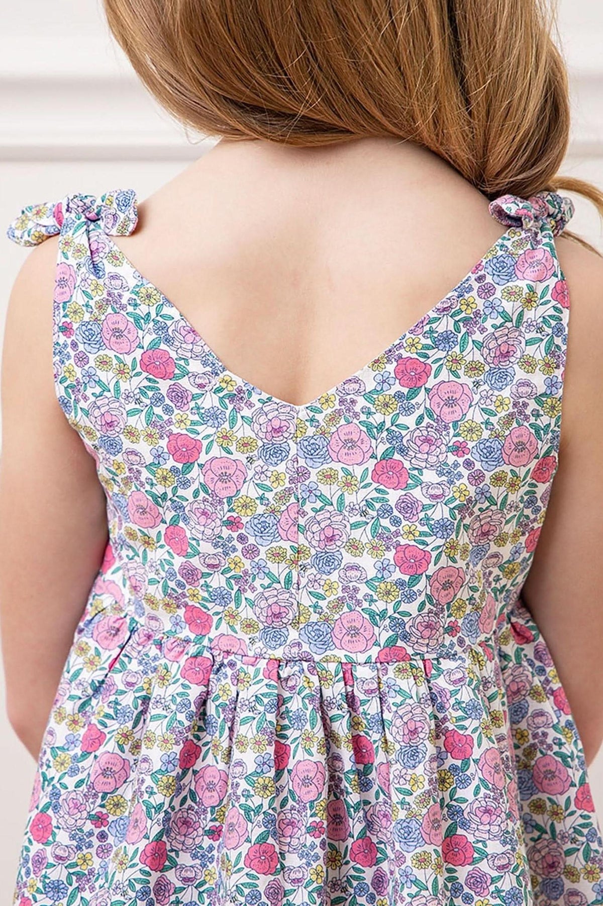 A close-up view of a girl wearing a floral linen dress featuring bow shoulder straps and a v-neckline. The colorful print includes pink and purple flowers on a white background.