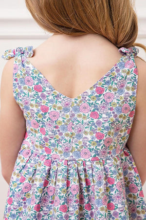 A close-up view of a girl wearing a floral linen dress featuring bow shoulder straps and a v-neckline. The colorful print includes pink and purple flowers on a white background.