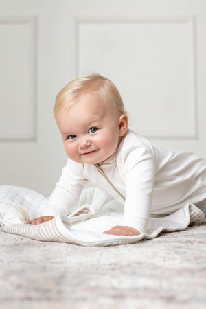 A smiling baby crawls on a soft, light-colored quilted blanket, wearing a white long-sleeve romper. The setting features a neutral backdrop, emphasizing the babys playful expression and cozy outfit.