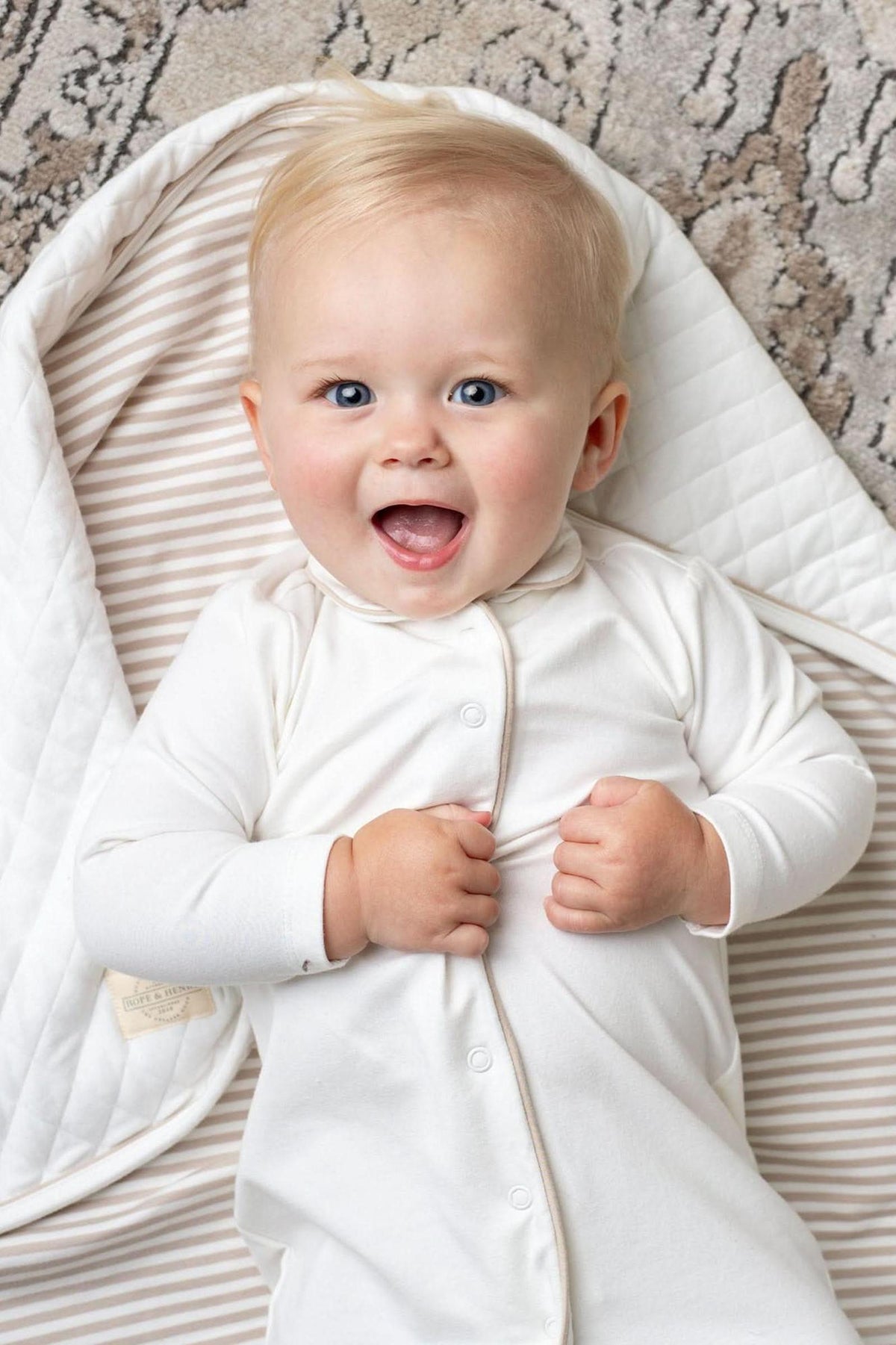 A joyful baby with blonde hair smiles while lying on a quilted, striped blanket. The baby is dressed in a soft, white long-sleeve romper, showcasing comfort and style.