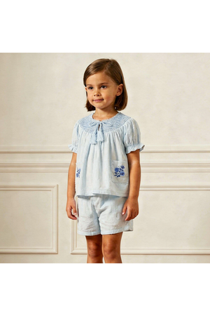 A young child models a light blue organic smocked top with short sleeves and matching shorts, featuring delicate embroidery. The soft, breathable fabric is perfect for warm, sunny days.