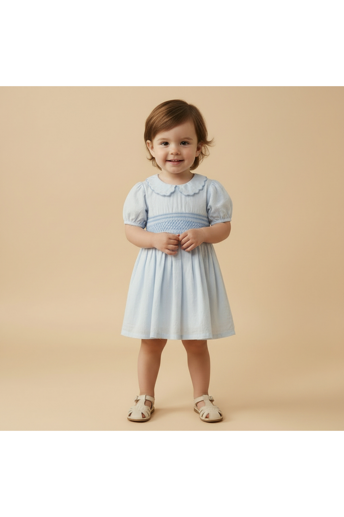 A toddler smiles while wearing a light blue organic cotton smocked collar dress with puff sleeves. The dress features a classic collar and is set against a soft beige background.