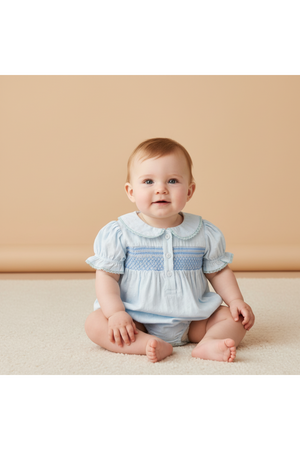 A baby sits on a soft, light-colored surface, wearing a light blue smocked romper with a classic collar and wooden buttons, showcasing a playful and timeless style.