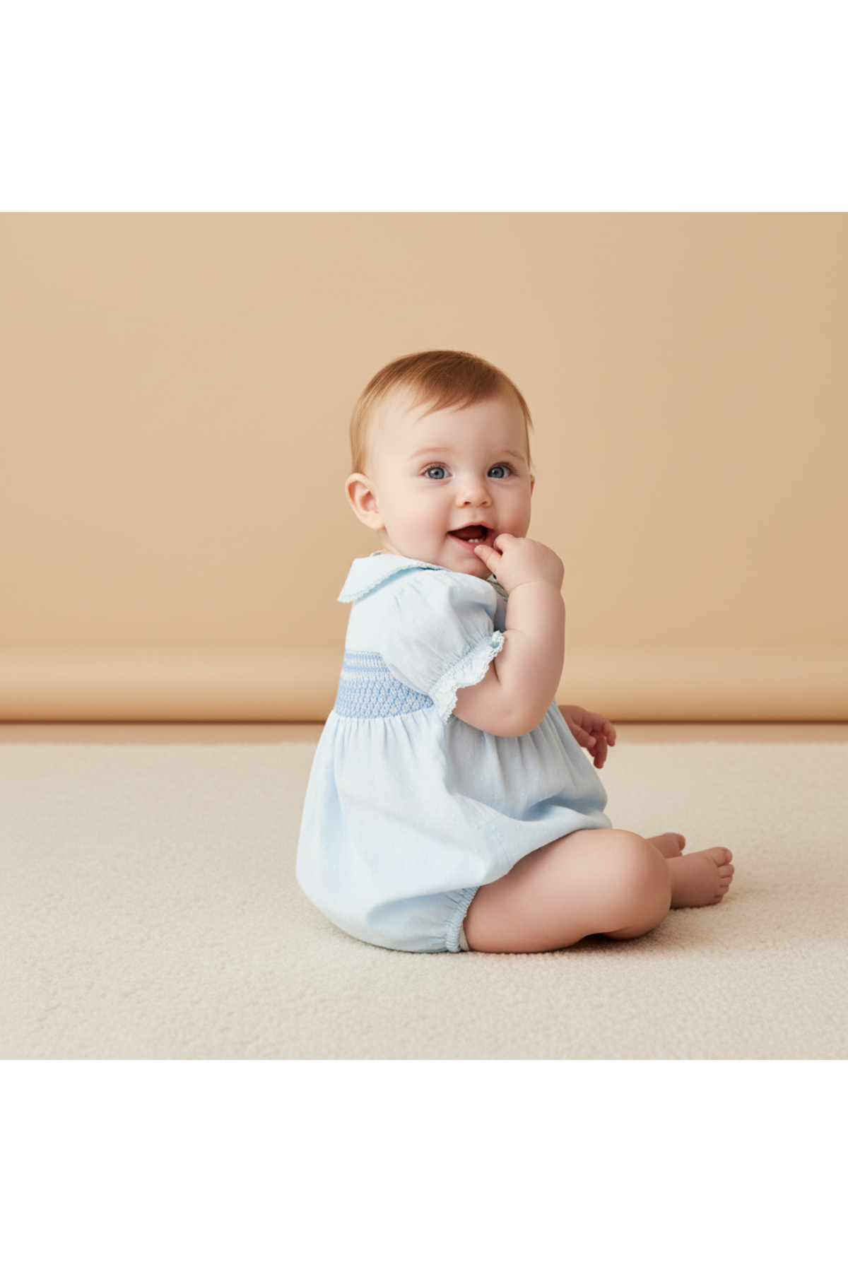 A baby sits on a soft, light-colored surface, wearing a light blue smocked romper with a collared neckline. The romper features a textured design and is made from organic cotton.