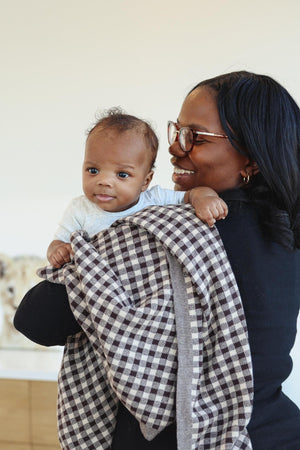 A baby held by a smiling adult, wrapped in a checkered knit blanket, in a cozy indoor setting.