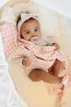 A baby with a pink pacifier and bow headband holds a pink checkered blanket in a cozy setting.