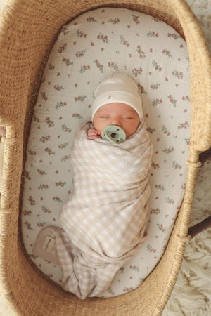 Newborn in a beige checkered swaddle, lying in a wicker basket with floral-patterned lining, wearing a white hat.